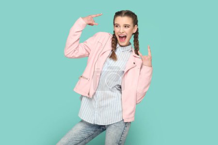 Téléchargez les photos : Portrait d'une adolescente extrêmement heureuse et excitée avec des tresses portant une veste rose montrant le rock and roll, s'amusant au festival. Studio intérieur tourné isolé sur fond vert. - en image libre de droit