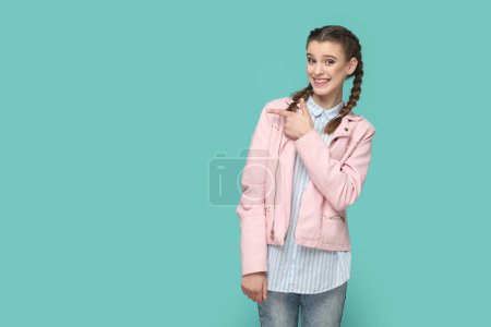 Foto de Retrato de la alegre joven adolescente encantadora con trenzas con chaqueta rosa que indica en la maqueta, espacio de copia para la publicidad. Estudio interior plano aislado sobre fondo verde. - Imagen libre de derechos