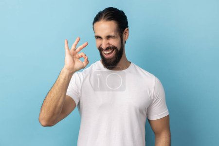 Téléchargez les photos : Portrait d'un homme heureux satisfait avec la barbe portant un T-shirt blanc debout, regardant la caméra montrant le geste Ok signe, exprimant des émotions positives. Studio intérieur tourné isolé sur fond bleu. - en image libre de droit