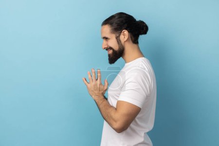 Photo for Side view of devious man with beard wearing white T-shirt smirking and conspiring cunning sly plan, pondering tricky clever idea in mind, wants to cheat. Indoor studio shot isolated on blue background - Royalty Free Image