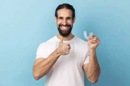 Photo for Portrait of man with beard wearing white T-shirt holding dental aligner retainer, dental clinic for beautiful teeth, showing thumb up. Indoor studio shot isolated on blue background. - Royalty Free Image