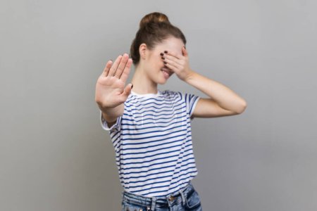 Photo for Portrait of afraid beautiful woman wearing striped T-shirt closing eyes with palm and showing stop hand gesture, turning face, does not want to see. Indoor studio shot isolated on gray background. - Royalty Free Image
