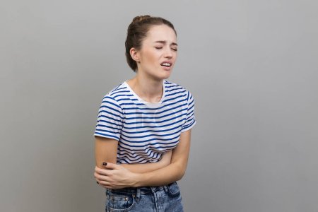 Photo for Portrait of unhealthy sick woman wearing striped T-shirt standing and touching her painful belly, frowning face from terrible pain. Indoor studio shot isolated on gray background. - Royalty Free Image