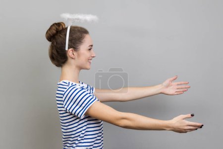 Photo for Side view of woman wearing striped T-shirt and with nimbus over her head stretching arms with kind friendly smile, going to embrace, share love. Indoor studio shot isolated on gray background - Royalty Free Image