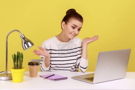 Téléchargez les photos : Femme incertaine employée de bureau en chemise rayée haussant les épaules assise sur un ordinateur portable sur le lieu de travail, regardant un moniteur, ayant un appel vidéo. Studio intérieur tourné isolé sur fond jaune. - en image libre de droit