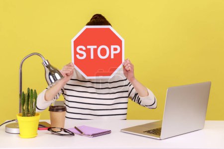 Photo for Woman office worker sitting at workplace hiding face behind red stop traffic sign avoiding conflicts, afraid of workplace bullying. Indoor studio studio shot isolated on yellow background. - Royalty Free Image