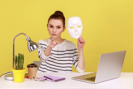 Photo for Serious woman office worker holding white face mask and pointing finger at camera, blaming you for hiding identity, duplicity. Indoor studio studio shot isolated on yellow background. - Royalty Free Image