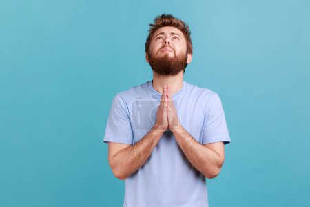Photo for Portrait of hopeless desperate bearded man folding hands in pray looking up, communicating with god, asking for forgiveness and blessing. Indoor studio shot isolated on blue background. - Royalty Free Image
