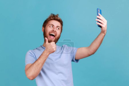 Foto de Retrato de un hombre barbudo sonriente positivo mostrando pulgares hacia el teléfono inteligente, comunicándose por videollamada, tomando selfie y haciendo gestos como. Estudio interior plano aislado sobre fondo azul. - Imagen libre de derechos