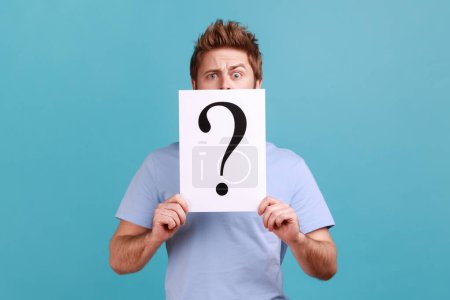 Photo for Portrait of puzzled bearded man covering half of face with white paper with question mark, finding smart solution, asking for advice. Indoor studio shot isolated on blue background. - Royalty Free Image