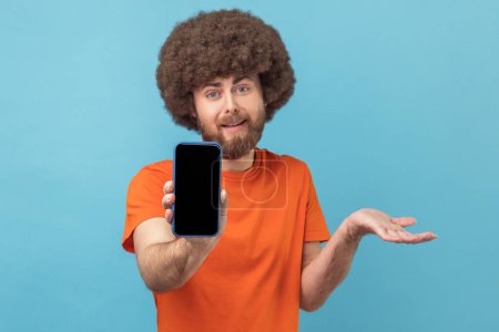 Photo for Portrait of confused man with Afro hairstyle wearing orange T-shirt showing cell phone with blank black screen, presenting area for advertisement. Indoor studio shot isolated on blue background. - Royalty Free Image