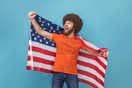 Téléchargez les photos : Portrait d'homme à la coiffure afro en T-shirt portant un énorme drapeau américain et se réjouissant tout en célébrant la fête nationale, détournant les yeux et criant. Studio intérieur tourné isolé sur fond bleu. - en image libre de droit