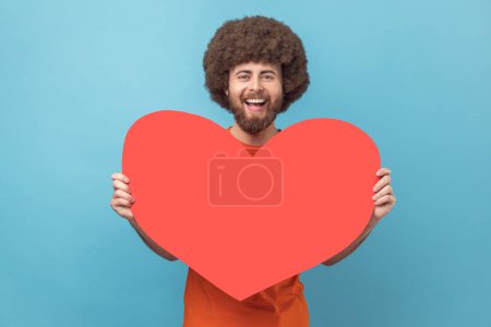 Photo for Portrait of man with Afro hairstyle wearing orange T-shirt holding big red heart, expressing positive romantic emotions, looking at camera with smile. Indoor studio shot isolated on blue background. - Royalty Free Image