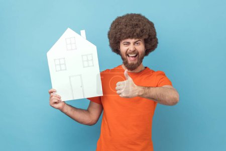 Téléchargez les photos : Portrait d'un homme positif excité avec une coiffure afro portant un T-shirt orange tenant une maison en papier, agence immobilière, montrant le pouce en l'air. Studio intérieur tourné isolé sur fond bleu. - en image libre de droit
