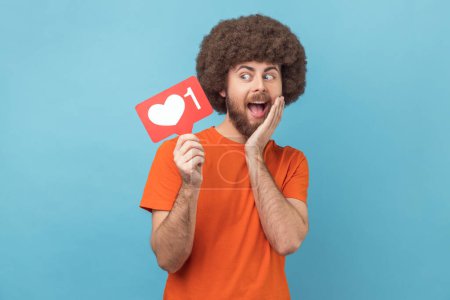 Photo for Portrait of cute surprised man with Afro hairstyle wearing orange T-shirt holding social media heart Like button, emoji counter, follower notification. Indoor studio shot isolated on blue background. - Royalty Free Image