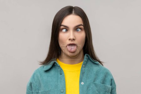 Photo for Portrait of dark haired woman sticking out tongue and looking at camera with crossed eyes, teasing with naughty expression, disobedient behavior. Indoor studio shot isolated on gray background. - Royalty Free Image
