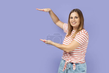 Foto de Retrato de una mujer rubia sonriente satisfecha con una camiseta a rayas que presenta el área entre las manos para publicidad, mostrando un tamaño enorme. Estudio interior plano aislado sobre fondo púrpura. - Imagen libre de derechos