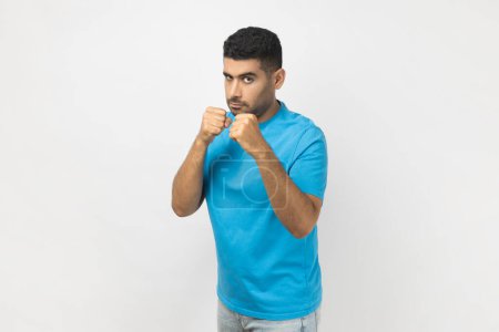 Photo for Portrait of confident serious unshaven man wearing blue T- shirt standing with clenched fists, being ready to attack, arguing with somebody. Indoor studio shot isolated on gray background. - Royalty Free Image