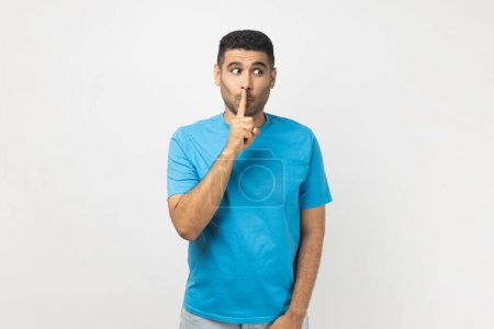 Photo for Portrait of funny positive unshaven man wearing blue T- shirt standing looking away, keeps finger near lips, promise to keep secret. Indoor studio shot isolated on gray background. - Royalty Free Image