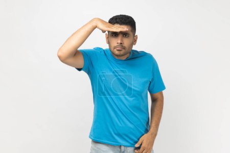 Photo for Portrait of unshaven man wearing blue T- shirt standing keeps hand near forehead, looks far away, searches something on horizon. Indoor studio shot isolated on gray background. - Royalty Free Image