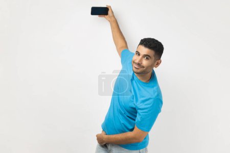 Téléchargez les photos : Homme joyeux et souriant en T-shirt bleu tenant un téléphone intelligent avec un écran vide dans les mains, en direct ou ayant un appel vidéo, regardant la caméra. Studio intérieur tourné isolé sur fond gris. - en image libre de droit