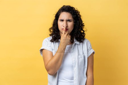 Photo for Portrait of woman with dark wavy hair looking with incredulous suspicious gaze and touching nose, gesturing you are liar, suspecting falsehood. Indoor studio shot isolated on yellow background. - Royalty Free Image