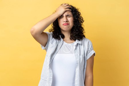 Photo for Portrait of woman with dark wavy hair standing with facepalm gesture, blaming herself, feeling sorrow regret because of bad memory. Indoor studio shot isolated on yellow background. - Royalty Free Image