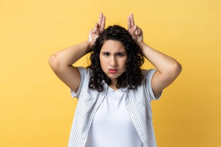 Photo for Portrait of strict serious woman with dark wavy hair showing bull horn gesture with fingers over head, looking hostile and threatening. Indoor studio shot isolated on yellow background. - Royalty Free Image