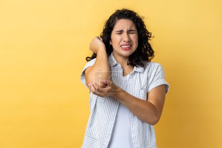 Portrait of woman with dark wavy hair having trauma after car accident, feels pain in elbow, trapped nerves, rheumatoid arthritis. Indoor studio shot isolated on yellow background.