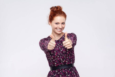 Photo for Portrait of smiling happy satisfied ginger woman wearing dress standing showing thumbs up, recommend excellent service. Indoor studio shot isolated on gray background. - Royalty Free Image