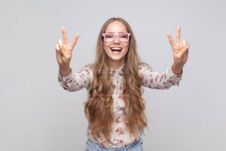 Photo for Belief in winning. Smiling pretty woman in glasses with wavy blond hair showing v sign with fingers, peace or victory gesture, rejoicing success. Indoor studio shot isolated on gray background. - Royalty Free Image