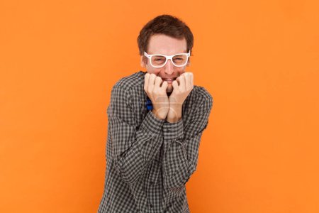 Photo for Anxious puzzled man nerd bites finger nails stands insecure and nervous, feels uncomfortable, wearing shirt with blue bow tie and white glasses. Indoor studio shot isolated on orange background. - Royalty Free Image