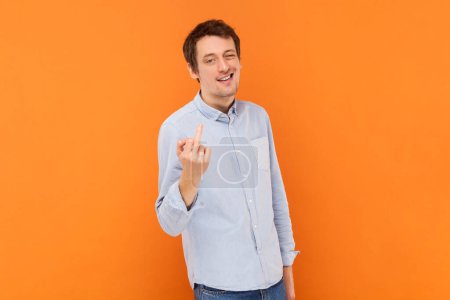 Photo for Portrait of serious vulgar selfish man showing middle finger, impolite gesture, disrespects someone, looks rude at camera, wearing light blue shirt. Indoor studio shot isolated on orange background. - Royalty Free Image