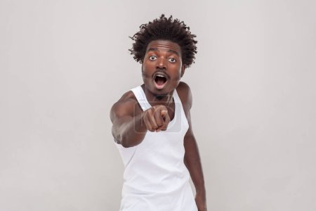 Photo for Omg look there. Shocked man with Afro hairstyle points index finger at camera with great surprise, holds breath indicates forward wearing white T-shirt. Indoor studio shot isolated on gray background. - Royalty Free Image