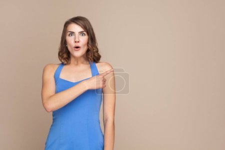 Photo for Portrait of surprised woman with wavy hair showing something aside at blank wall, being astonished, advertisement area, wearing blue dress. Indoor studio shot isolated on light brown background. - Royalty Free Image