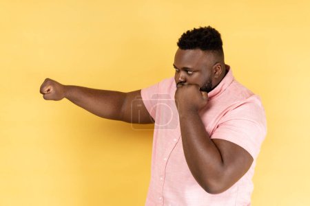 Photo for I'll hit you. Side view of bearded man wearing pink shirt standing with raised fists boxing gesture ready to punch, ready to struggle, fighting spirit. Indoor studio shot isolated on yellow background - Royalty Free Image