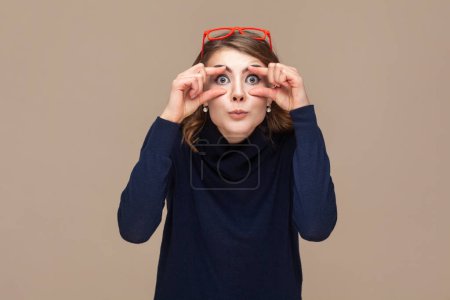 Photo for Portrait of funny surprised astonished woman with wavy hair in red red glasses on her head opens her eyes with fingers to see better. Indoor studio shot isolated on light brown background. - Royalty Free Image