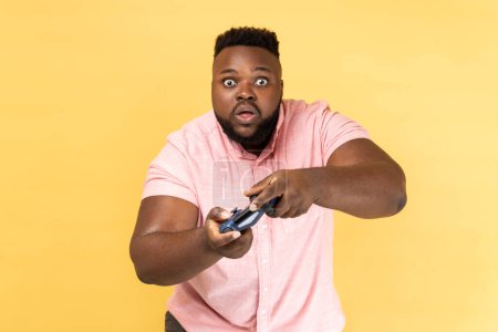 Photo for Portrait of shocked surprised bearded man wearing pink shirt playing games, holding joypad, looking at camera with big eyes and open mouth. Indoor studio shot isolated on yellow background. - Royalty Free Image