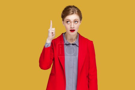 Photo for Portrait of excited amazed woman with red lips standing indicating finger upwards, having good excellent idea, wearing red jacket. Indoor studio shot isolated on yellow background. - Royalty Free Image