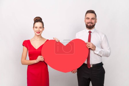 Photo for Portrait of smiling man in white shirt and woman in red dress standing together, holding big heart, celebrating Valentines Day. expressing love. Indoor studio shot isolated on gray background. - Royalty Free Image