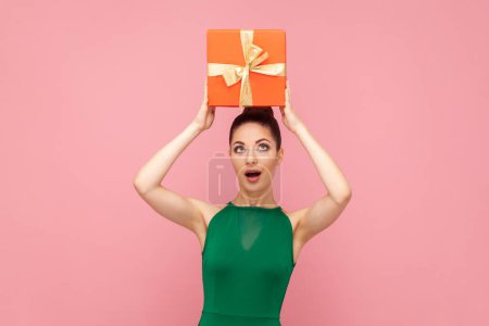 Téléchargez les photos : Portrait d'une femme étonnée étonnée surprise tenant une boîte cadeau rouge au-dessus de sa tête, excitée, portant une robe verte. Studio intérieur tourné isolé sur fond rose. - en image libre de droit