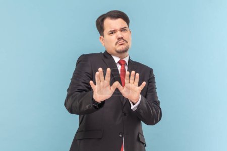 Photo for Portrait of frighten man with mustache standing raised arms, showing stop block gesture, being scared, wearing black suit with red tie. Indoor studio shot isolated on light blue background. - Royalty Free Image