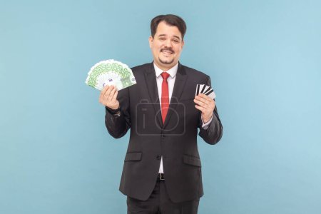 Photo for Satisfied rich man with mustache standing holding euro banknotes and credit cards in hands, being happy, wearing black suit with red tie. Indoor studio shot isolated on light blue background. - Royalty Free Image