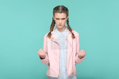 Photo for Portrait of angry aggressive attractive teenager girl with braids wearing pink jacket standing with clenched fits, arguing with somebody. Indoor studio shot isolated on green background. - Royalty Free Image
