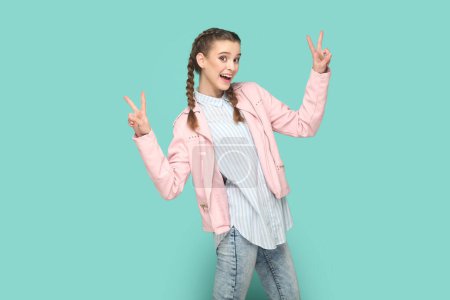 Photo for Portrait of excited optimistic cheerful teenager girl with braids wearing pink jacket showing v signs, being happy, being in high spirit. Indoor studio shot isolated on green background. - Royalty Free Image