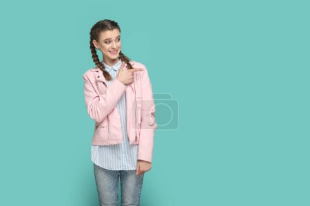 Foto de Retrato de la sonrisa positiva adolescente feliz con trenzas con chaqueta rosa señalando a un lado en el área de publicidad, espacio para la promoción. Estudio interior plano aislado sobre fondo verde. - Imagen libre de derechos