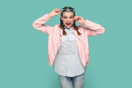 Excited amazed teenager girl with braids wearing pink jacket standing in two colorful optical spectacles, looking at camera with surprised expression. Indoor studio shot isolated on green background.