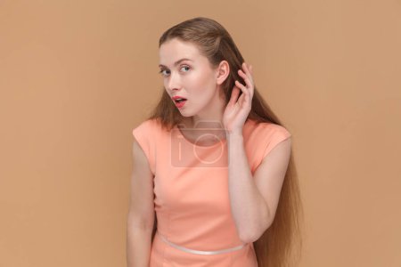 Photo for Portrait of attarctive lovely woman with long hair standing with hand near ear, listening to somebody whisper, wearing elegant dress. Indoor studio shot isolated on brown background. - Royalty Free Image