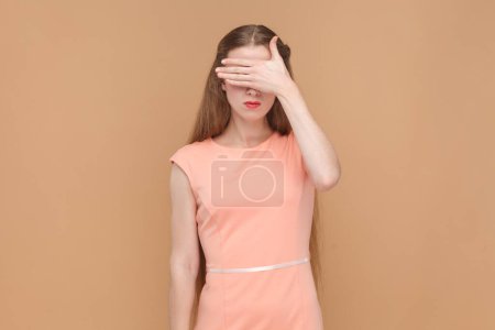 Photo for Portrait of serious sad woman with long hair standing and covering her eyes with her palms, doesn't want to see something shame, wearing elegant dress. Indoor studio shot isolated on brown background. - Royalty Free Image