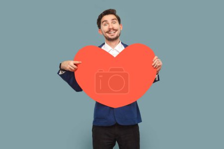 Photo for Portrait of handsome man with mustache standing holding pointing at big red heart, smiling toothily to camera, wearing white shirt and jacket. Indoor studio shot isolated on light blue background. - Royalty Free Image